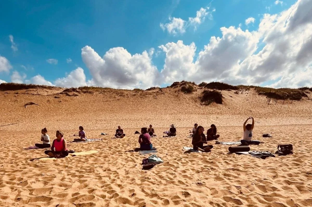 Groupe de personnes faisant du yoga sur la plage, assises sur des tapis, avec un grand dune et un ciel bleu avec des nuages en arrière-plan.