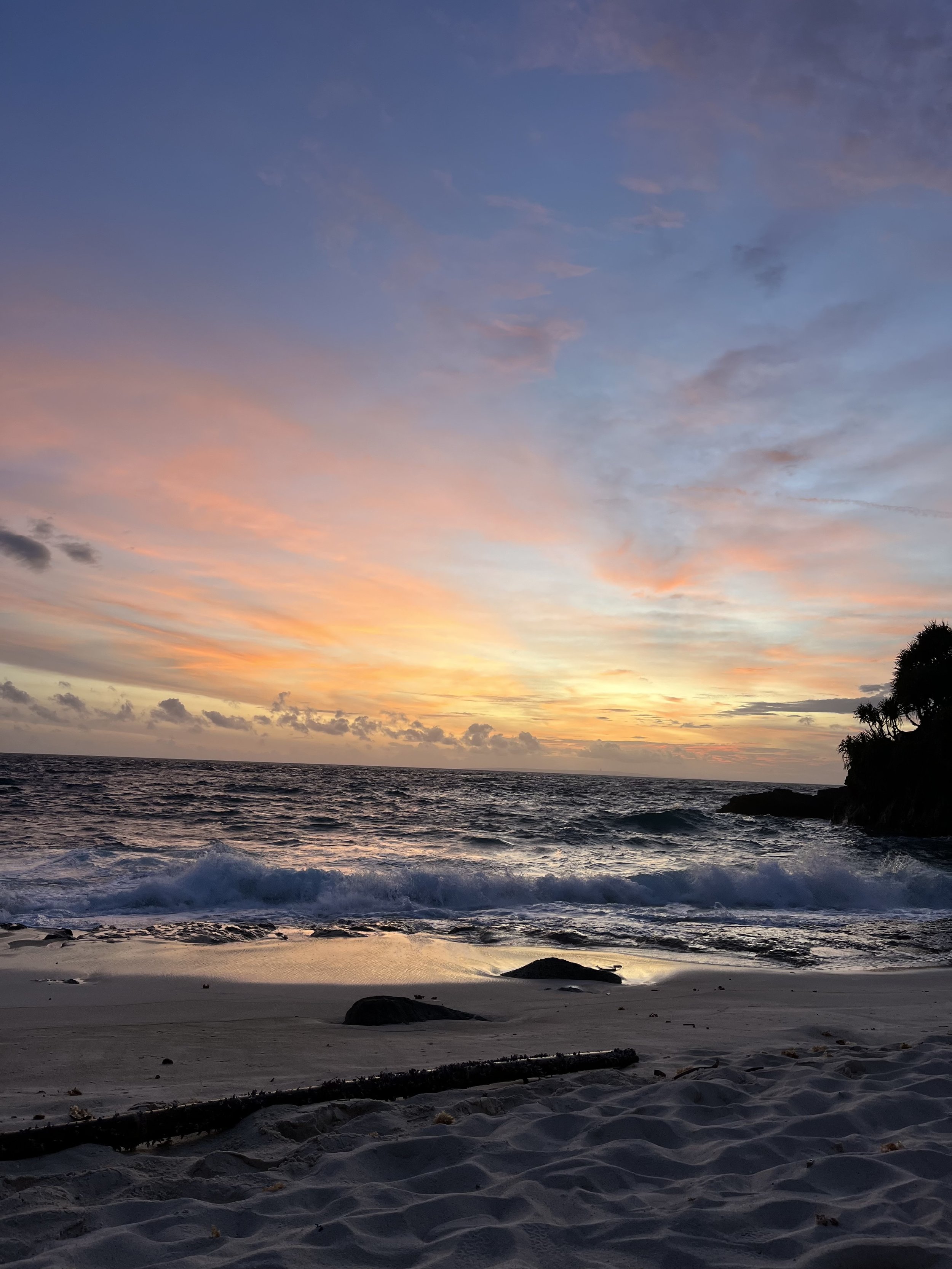 Coucher de soleil sur une plage, ciel coloré avec des nuances de rose, orange et bleu, vagues saccadées et sable avec quelques galets et débris sur la côte