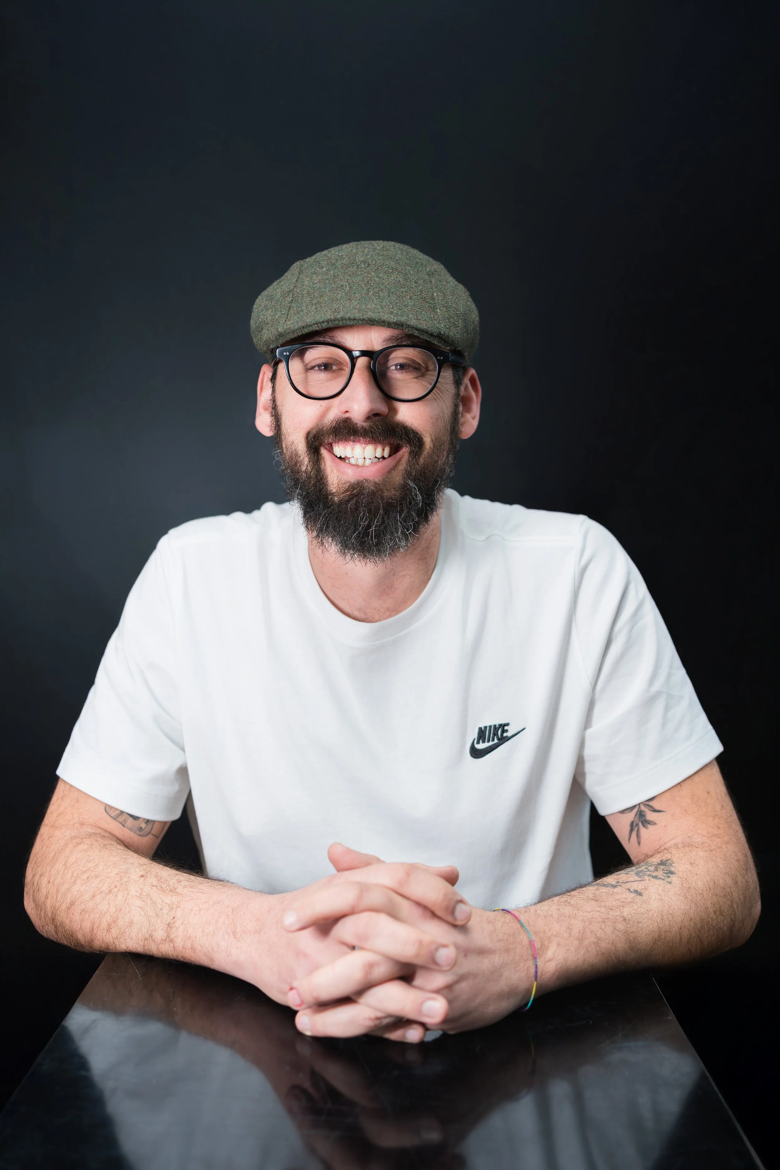 Uomo sorridente con barba, occhiali e cappello verde, seduto al tavolo in una stanza con sfondo nero, indossa una maglietta bianca Nike.