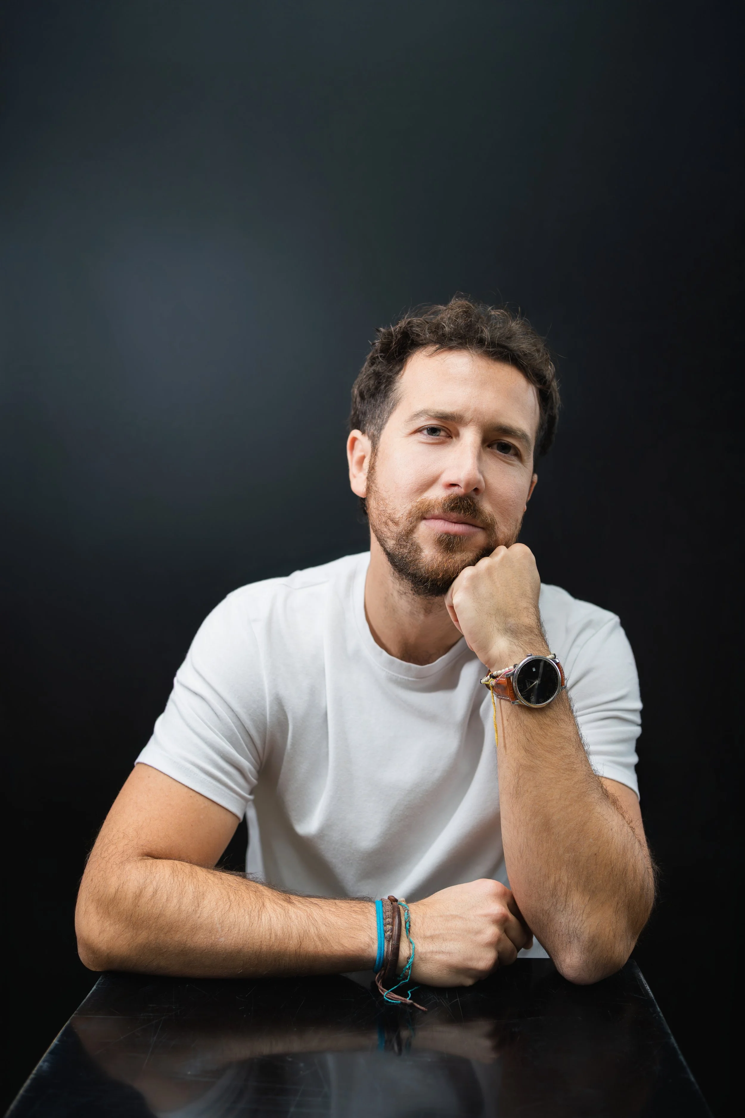Ritratto di un uomo con barba e capelli ricci, che indossa un orologio al polso, seduto davanti a una superficie nera, appoggiando il viso sulla mano, con sfondo nero.