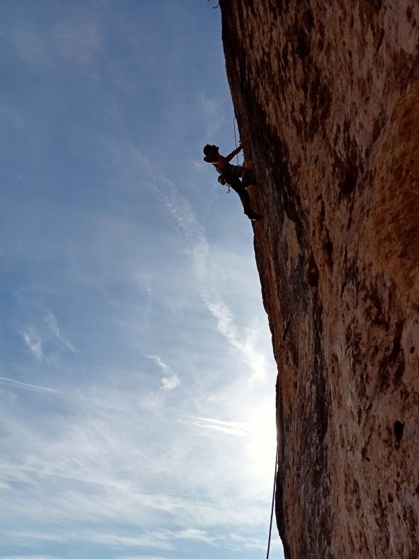 Une personne en train de faire de l’escalade sur une falaise abrupte brun-rouge, sous un ciel partiellement nuageux.