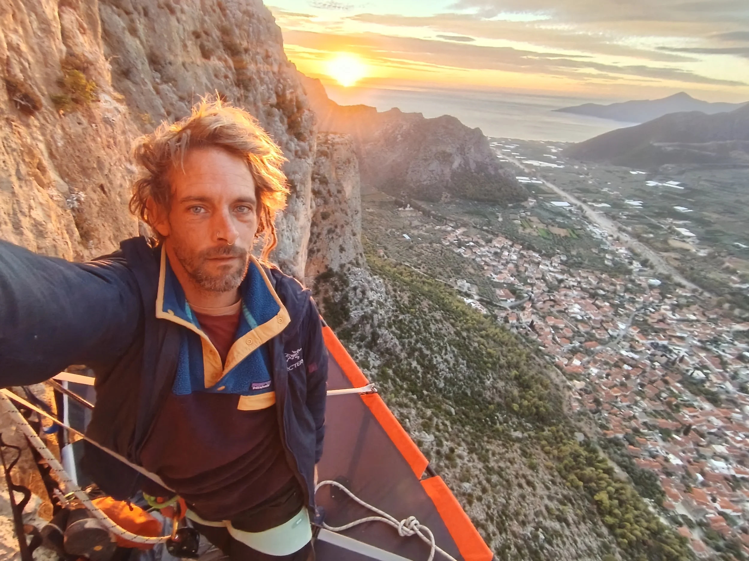 Un homme en equipement d'escalade pris en selfie sur une falaise au coucher du soleil, avec un paysage de rivière, champs et montagnes en arrière-plan.
