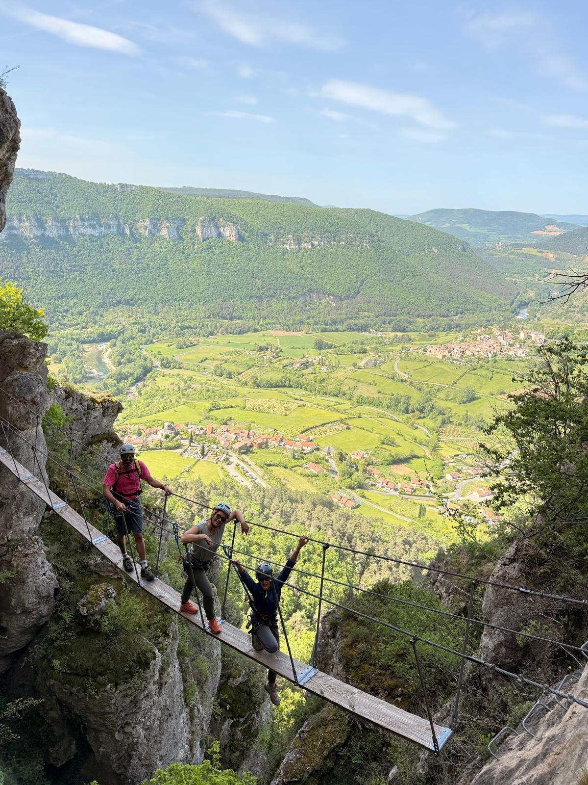 Quatre personnes portant des casques de sécurité traversant un pont suspendu en canyon, avec un paysage de montagnes et de champs en arrière-plan.