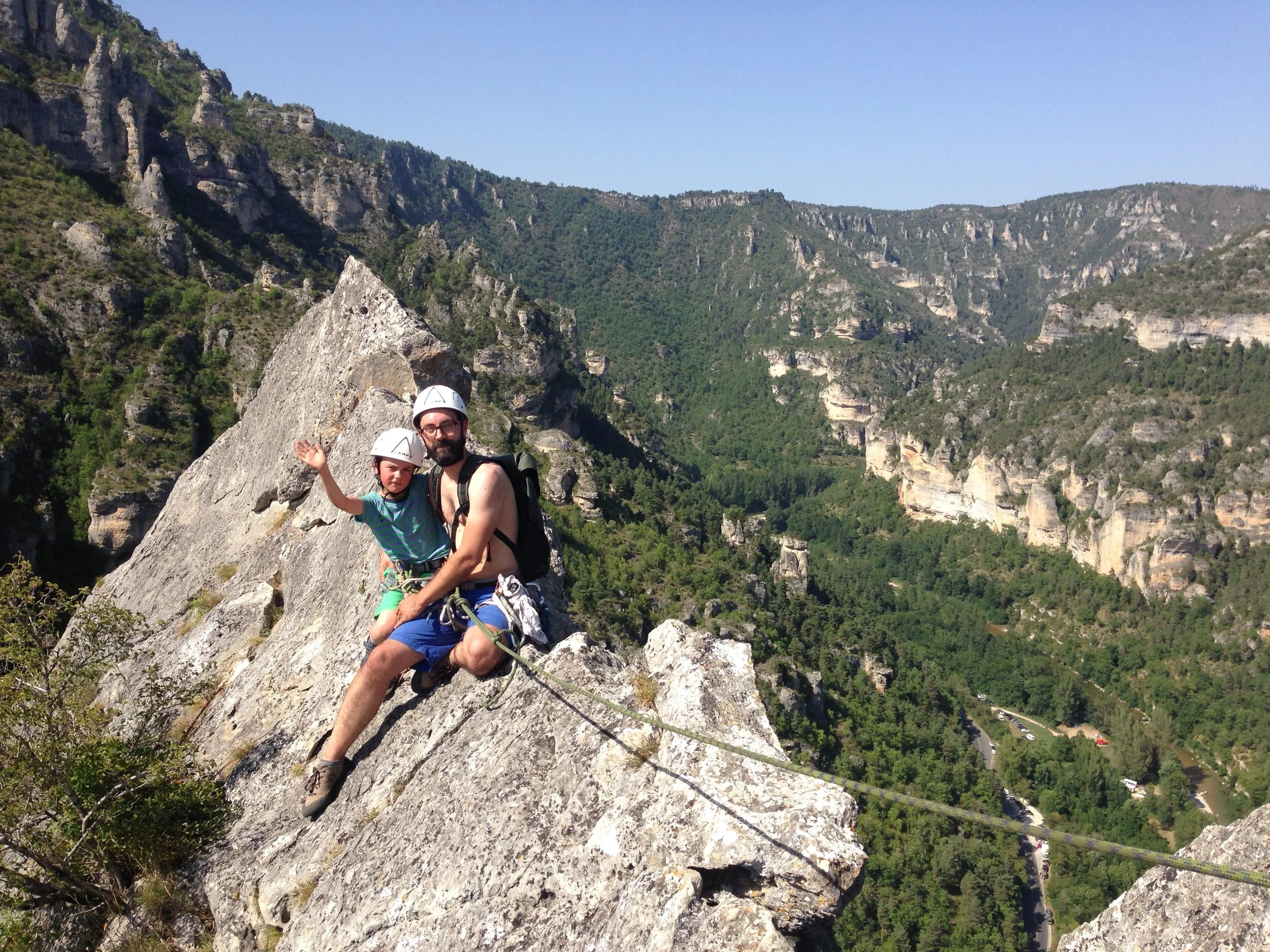Un homme et un jeune enfant, équipés de casques et de harnais, escaladent une falaise abrupte dans les Gorges du Tarn, avec une forêt verdoyante et une rivière en contrebas.
