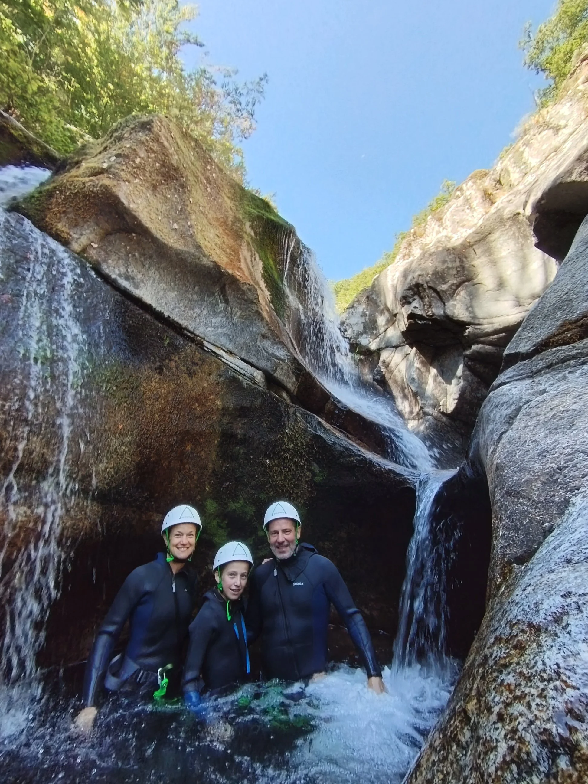 Groupe de personnes souriantes équipées de casques de protection, debout dans une rivière entourée de rochers, sous une cascade naturelle en forêt, ciel bleu en arrière-plan.