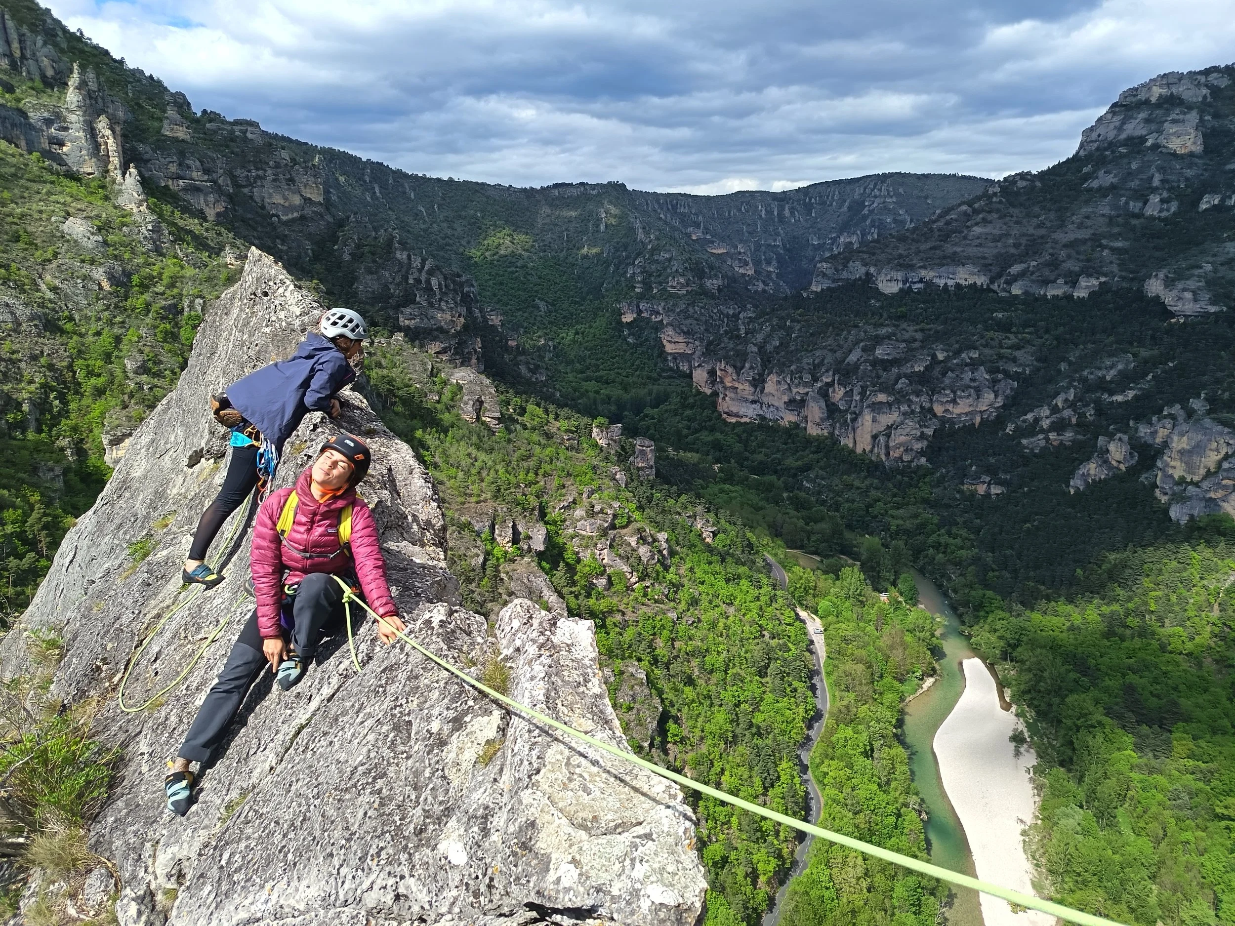 Deux personnes en escalade en haut d'une falaise rocheuse, avec un paysage de montagnes et une vallée avec un lac en contrebas, sous un ciel nuageux.
