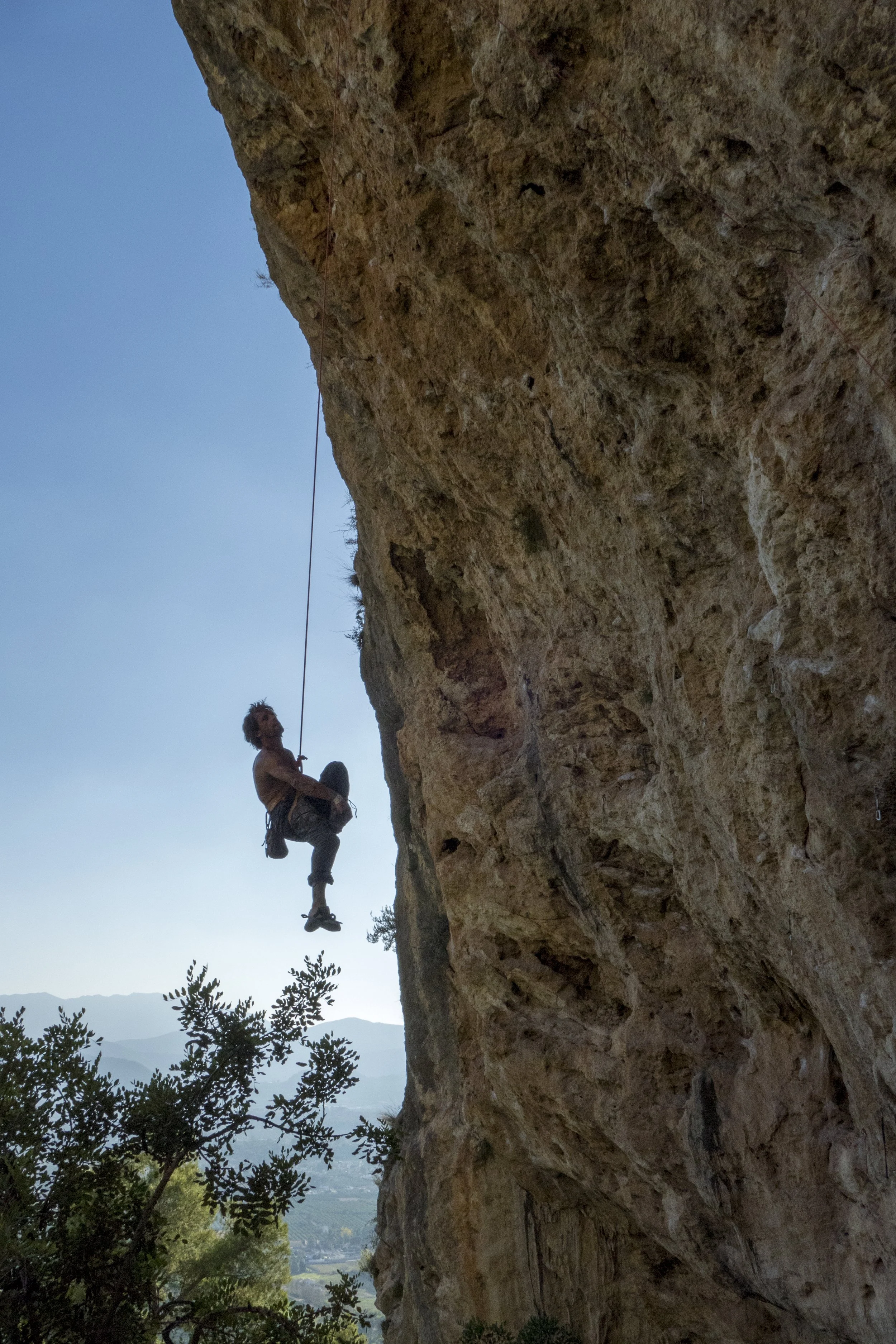Une personne escaladant une paroi rocheuse avec un harnais, suspendue à une corde, avec en arrière-plan des montagnes et des arbres.