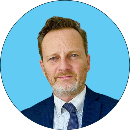 A very handsome man with gray hair and a beard wearing a suit and tie, looking at the camera against a blue background. Conveying trust, charm and hopeless optimism.
