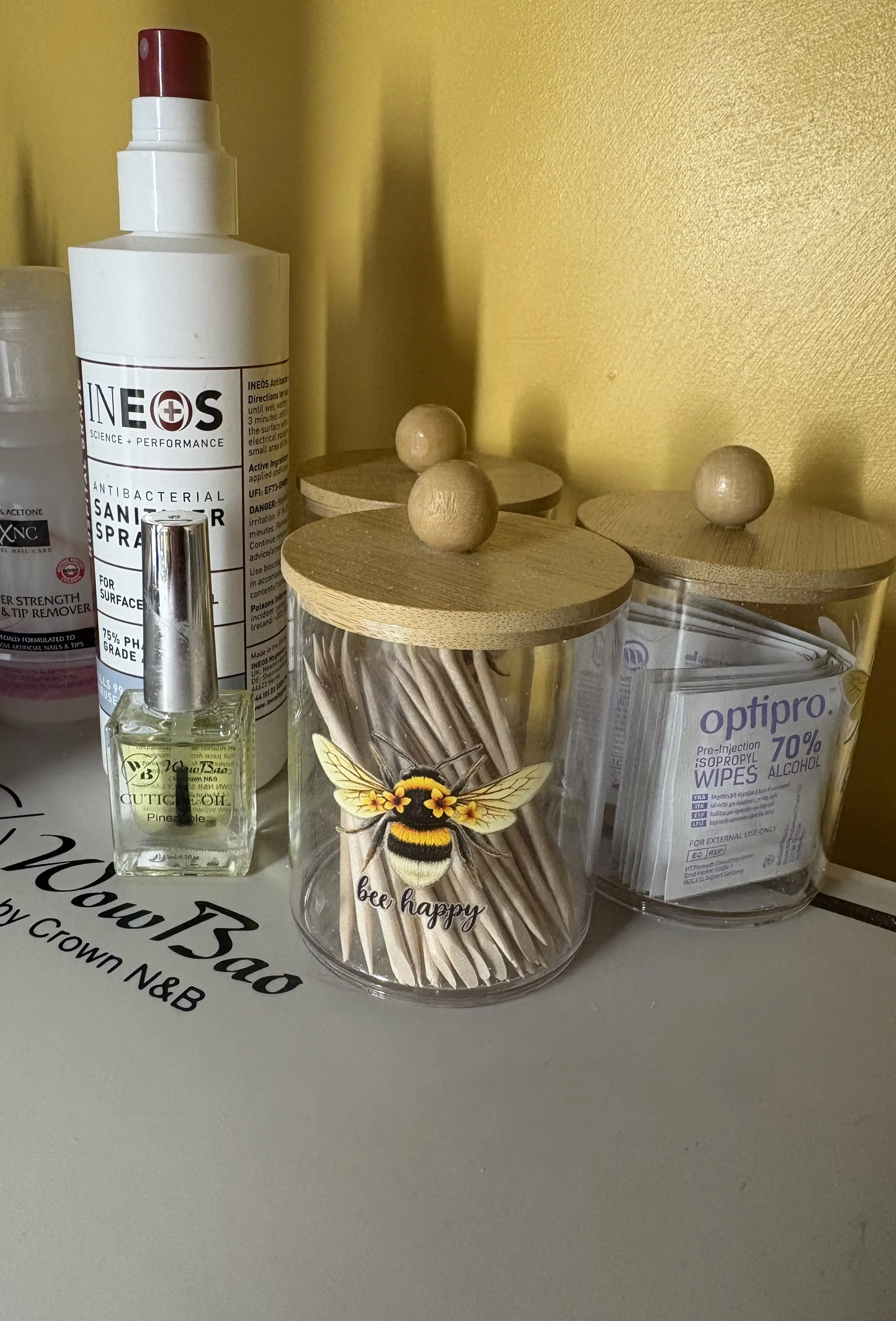 Collection of small containers and bottles on a surface against a yellow wall, including a sanitizer spray, a small bottle of cuticle oil with pine scent, and a container of Optipro alcohol wipes, with a decorative jar featuring a bee and the words 'bee happy' in the center.