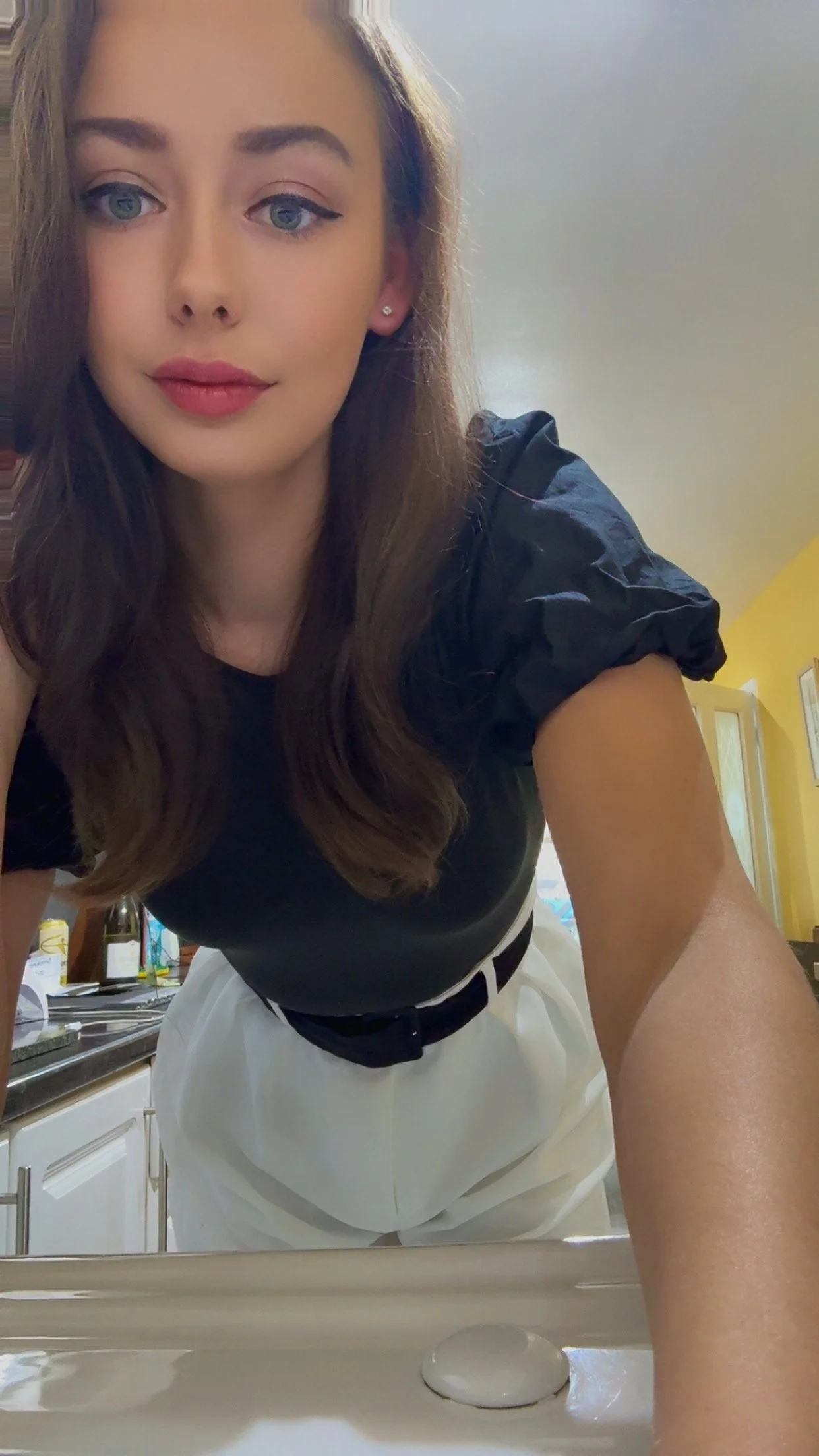 A young woman with long brown hair, wearing a black top with puffed sleeves and white pants, is leaning over a kitchen sink and looking into the camera. The background shows a kitchen with yellow walls and various bottles on the counter.