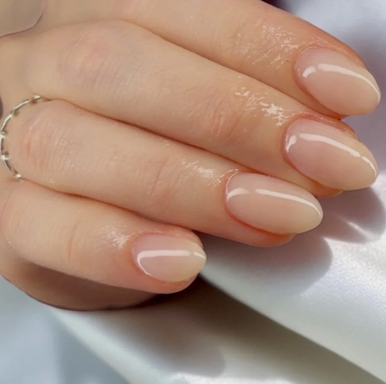 Close-up of neatly manicured fingernails with natural glossy nude polish, resting on a white surface.