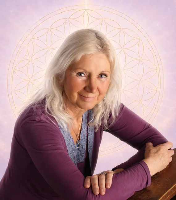 Dee Banton, author and spiritual teacherA mature  woman with long white  hair, wearing a purple top, sitting at a wooden table with her arms crossed, in front of a pastel-colored background with a golden geometric pattern resembling a flower of life.