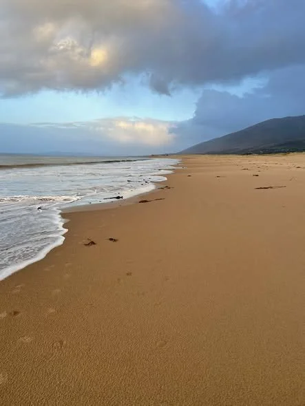 Empty sandy beach with footprints, ocean waves, and cloudy sky with mountains in the background.