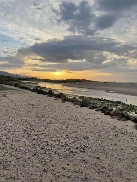 A sandy beach at sunset with a line of rocks and a cloudy sky over the ocean.