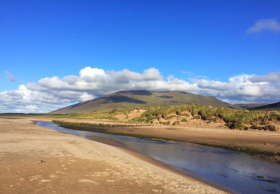 A scenic landscape featuring a small stream flowing through a sandy beach with green vegetation, mountains in the background, and a blue sky with scattered clouds.