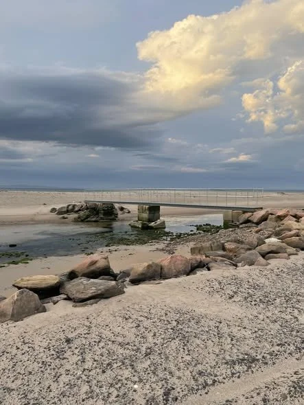A bridge over a sandy beach with rocks and water, under a partly cloudy sky.