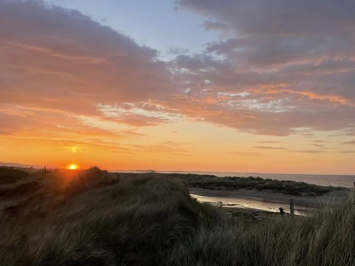 Sunset over a sandy beach with grassy dunes and water in the distance, with a partly cloudy sky.