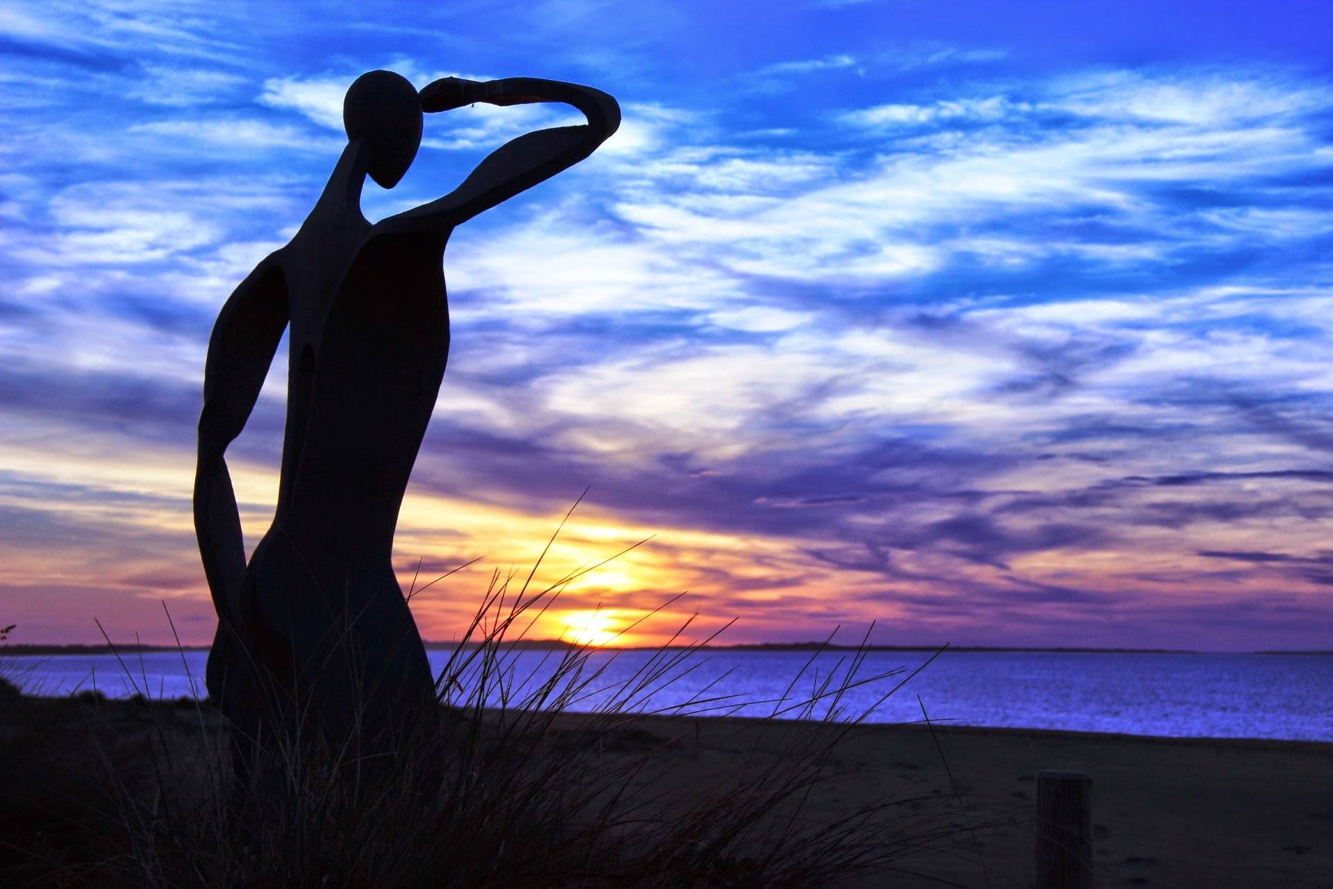 Silhouette of a hand forming a heart shape against a sunset sky with clouds over a body of water.