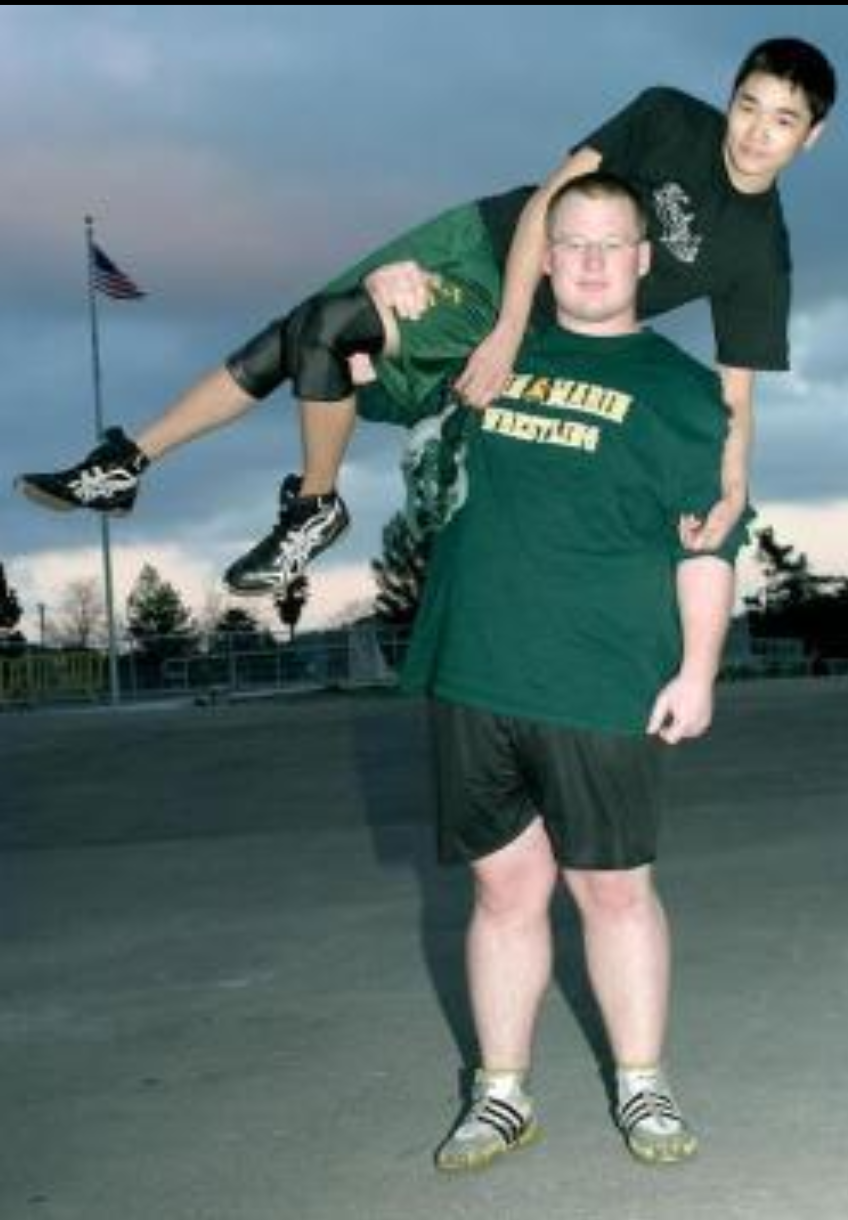 Two young men at an outdoor sports court, one holding the other horizontally; the man being held wears a black T-shirt, green shorts, and sneakers, while the man holding wears a green T-shirt, black shorts, and sandals. An American flag is visible in the background.