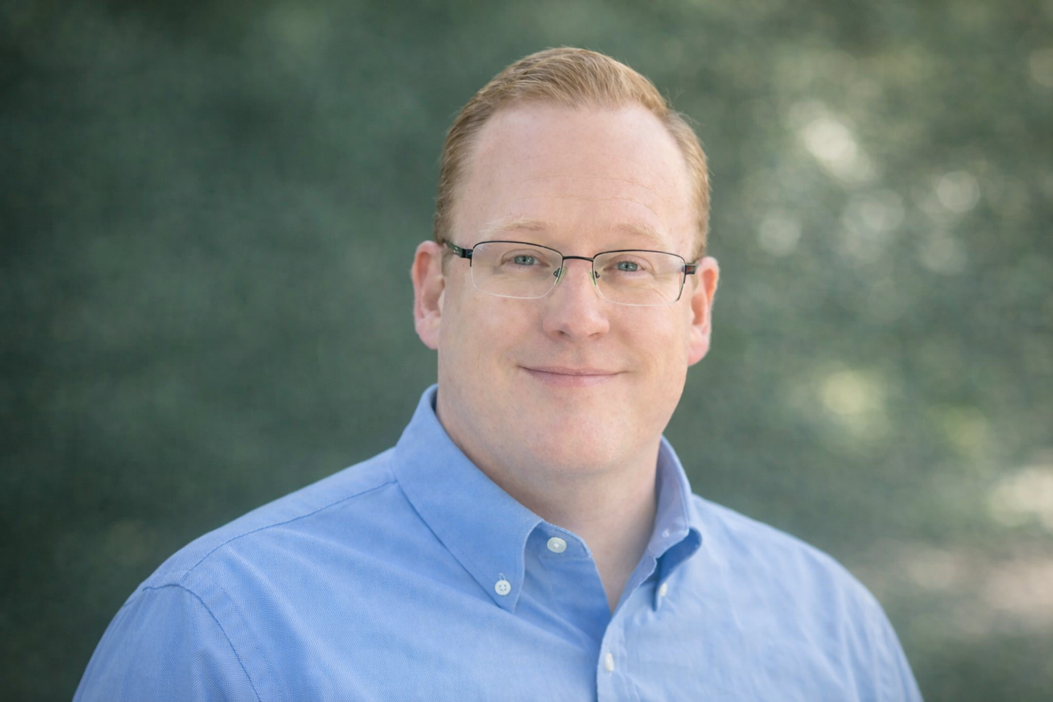 A man with red hair and glasses smiling outdoors, wearing a blue button-up shirt, with a blurred natural background.