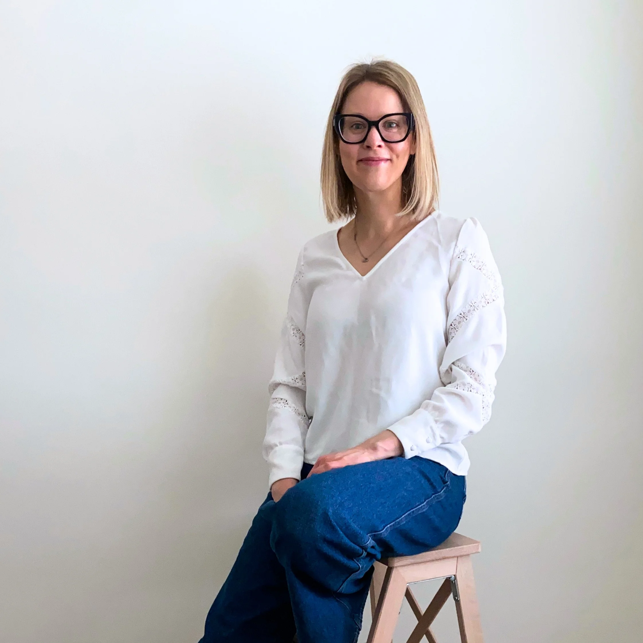 A woman with shoulder-length blonde hair, wearing glasses, a white long-sleeve blouse, and blue jeans, sitting on a wooden stool against a plain white wall.