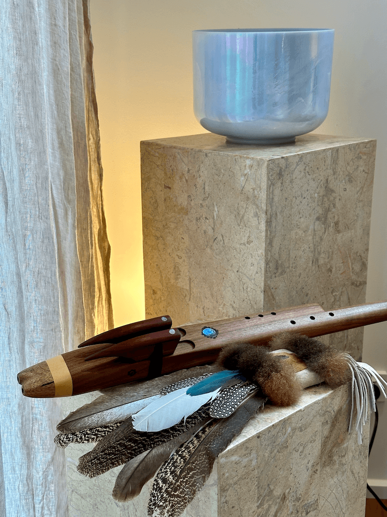 A wooden flute with feathers, fur, and beads decorated on it. The flute is resting on a stone pedestal, with a ceramic bowl on a higher stone pedestal in the background.