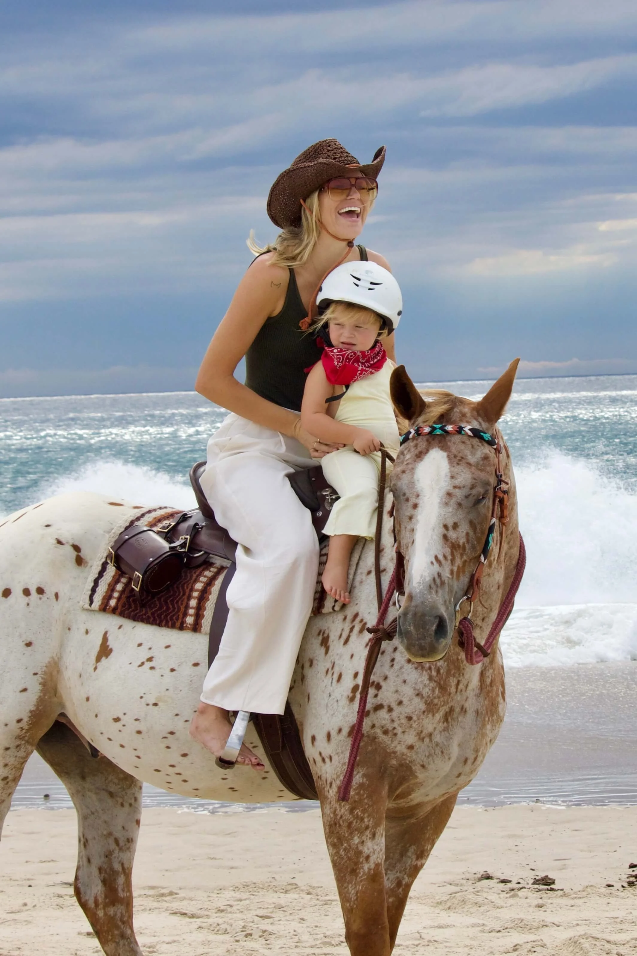 A woman and a young girl riding a spotted horse on a beach, with the ocean and sky in the background.