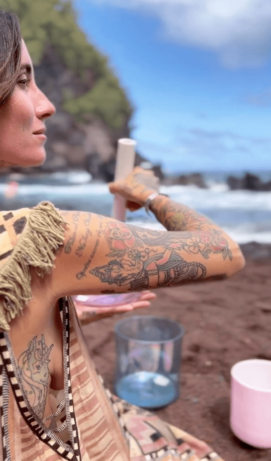 A woman with tattoos on her arm sitting on a beach, holding a white object, with a glass of water and cups on the sand in front of her, ocean waves and rocky cliffs in the background.