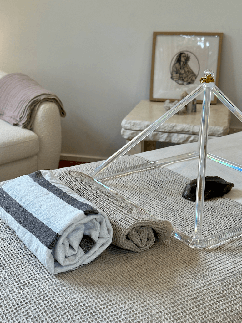 Stacked rolled towels, a glass pyramid display, and a small dark object on a textured beige surface in a softly lit room with a framed picture on the wall in the background.
