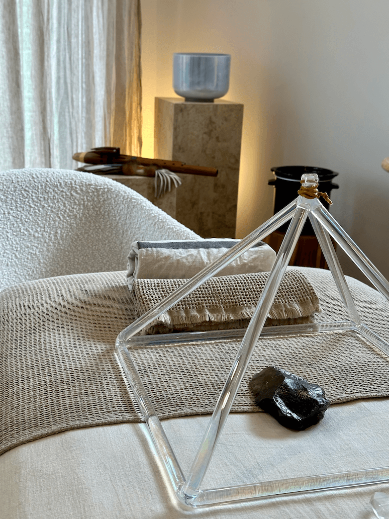 Close-up of a decorative room with a glass geometric sculpture, a dark rock, pillows on a bed, and a wooden chopstick on a bed's headboard with a beige throw blanket in a softly lit room.