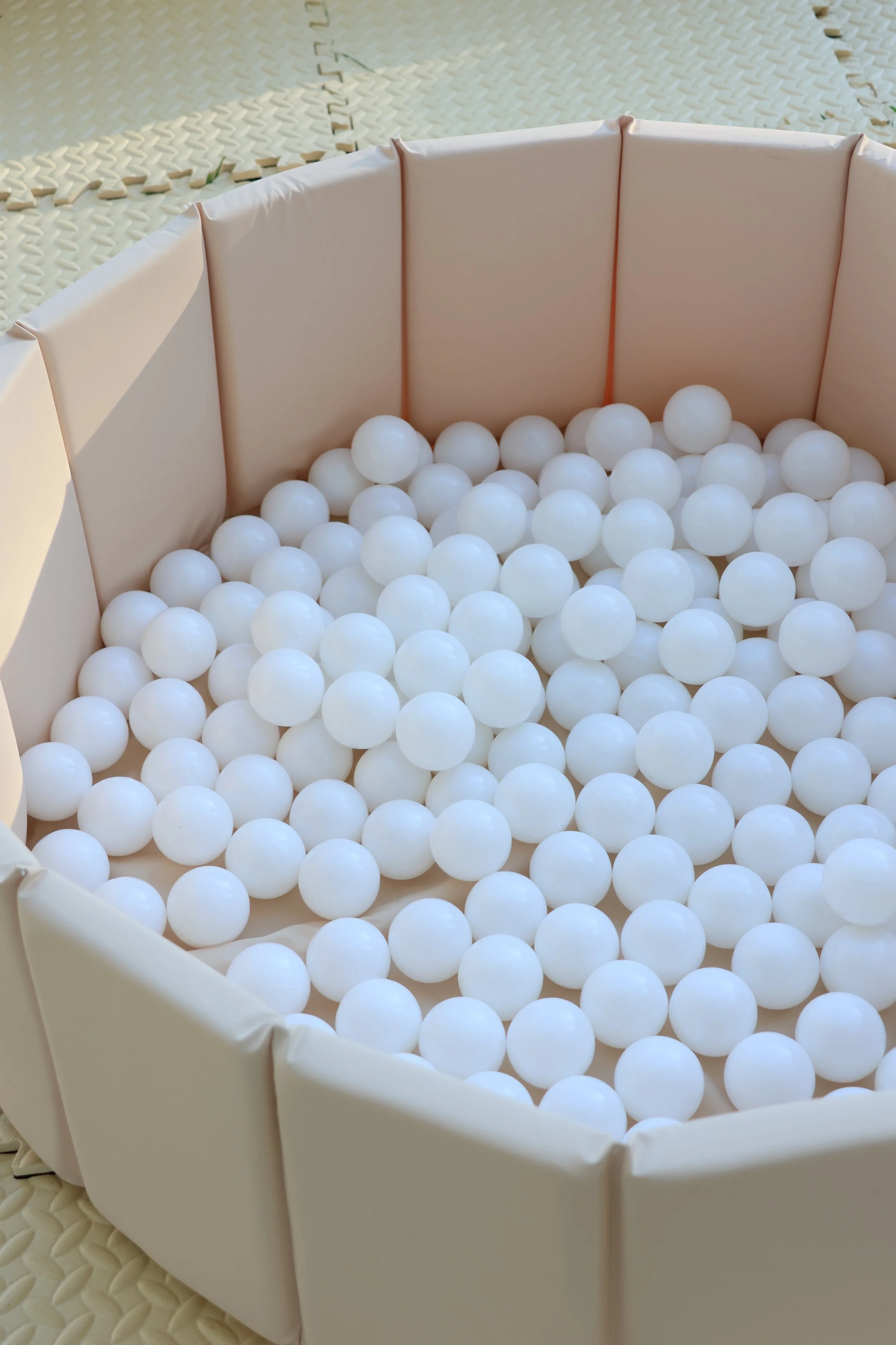 Empty ball pit with white plastic balls inside a beige cushioned enclosure, located on a foam floor covering.