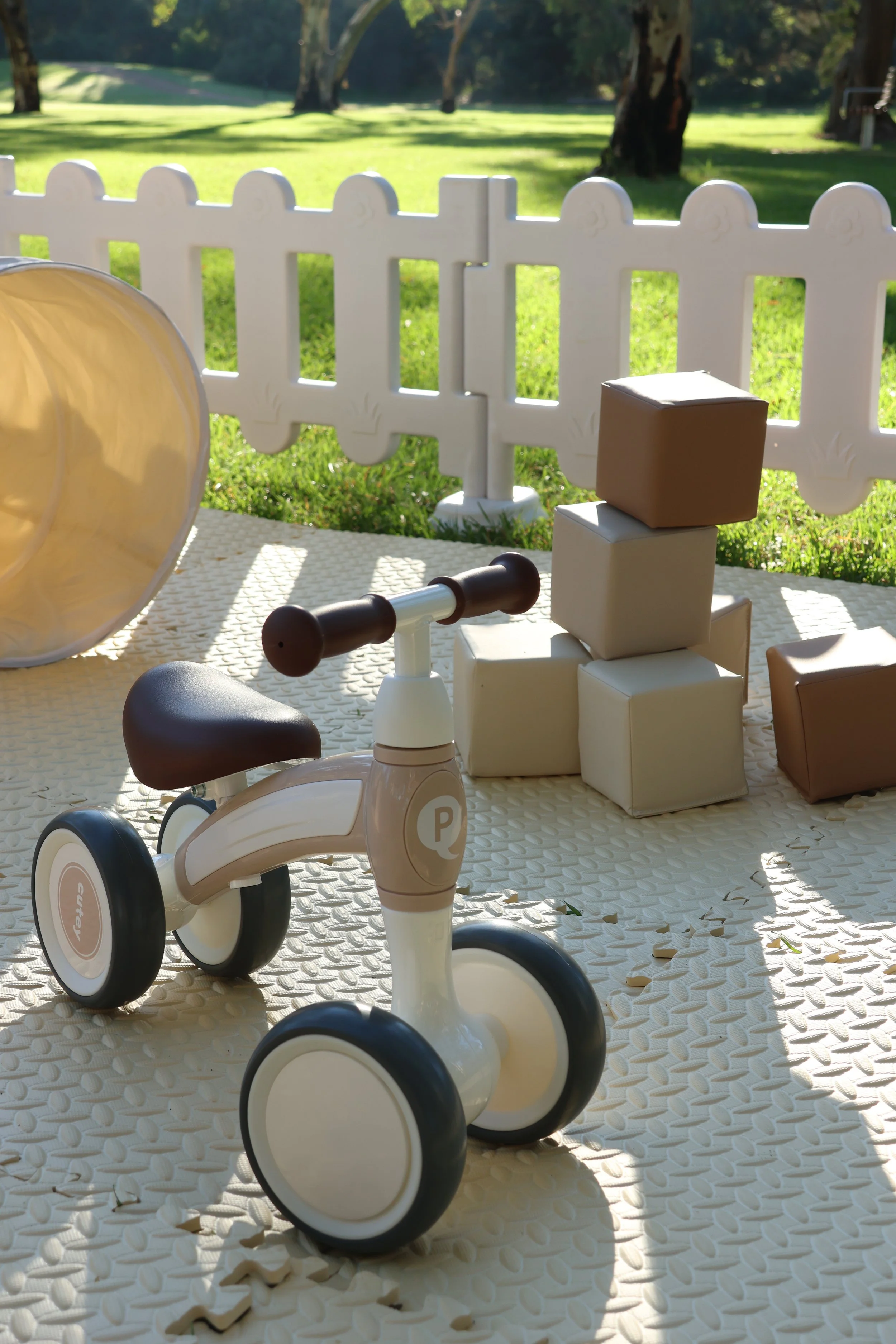 Children's toy bicycle and stacked cubical blocks on a textured surface outdoors, with a white picket fence and trees in the background.