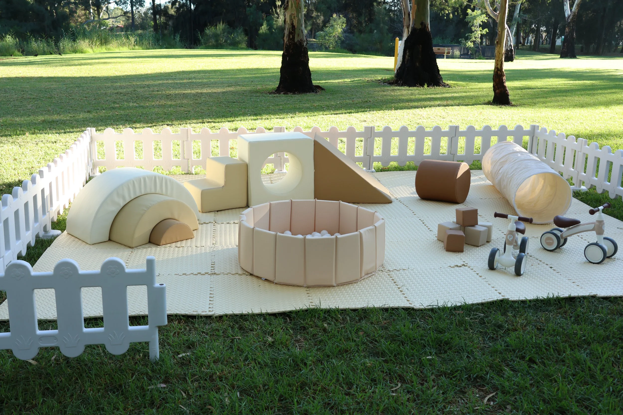 Outdoor play area with soft foam climbing structures, balls, toy blocks, a small slide, tricycle, and a tunnel, enclosed by a white picket fence on a grassy field with trees in the background.