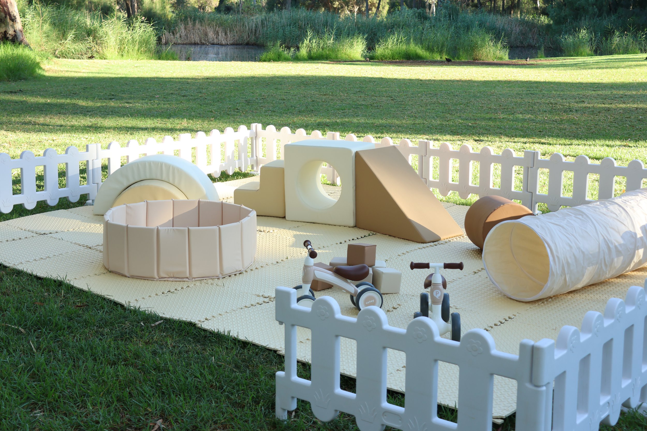 Outdoor play area with foam climbing structures, a padded tunnel, a ball pit, and toy bikes on a grassy lawn surrounded by a white picket fence.