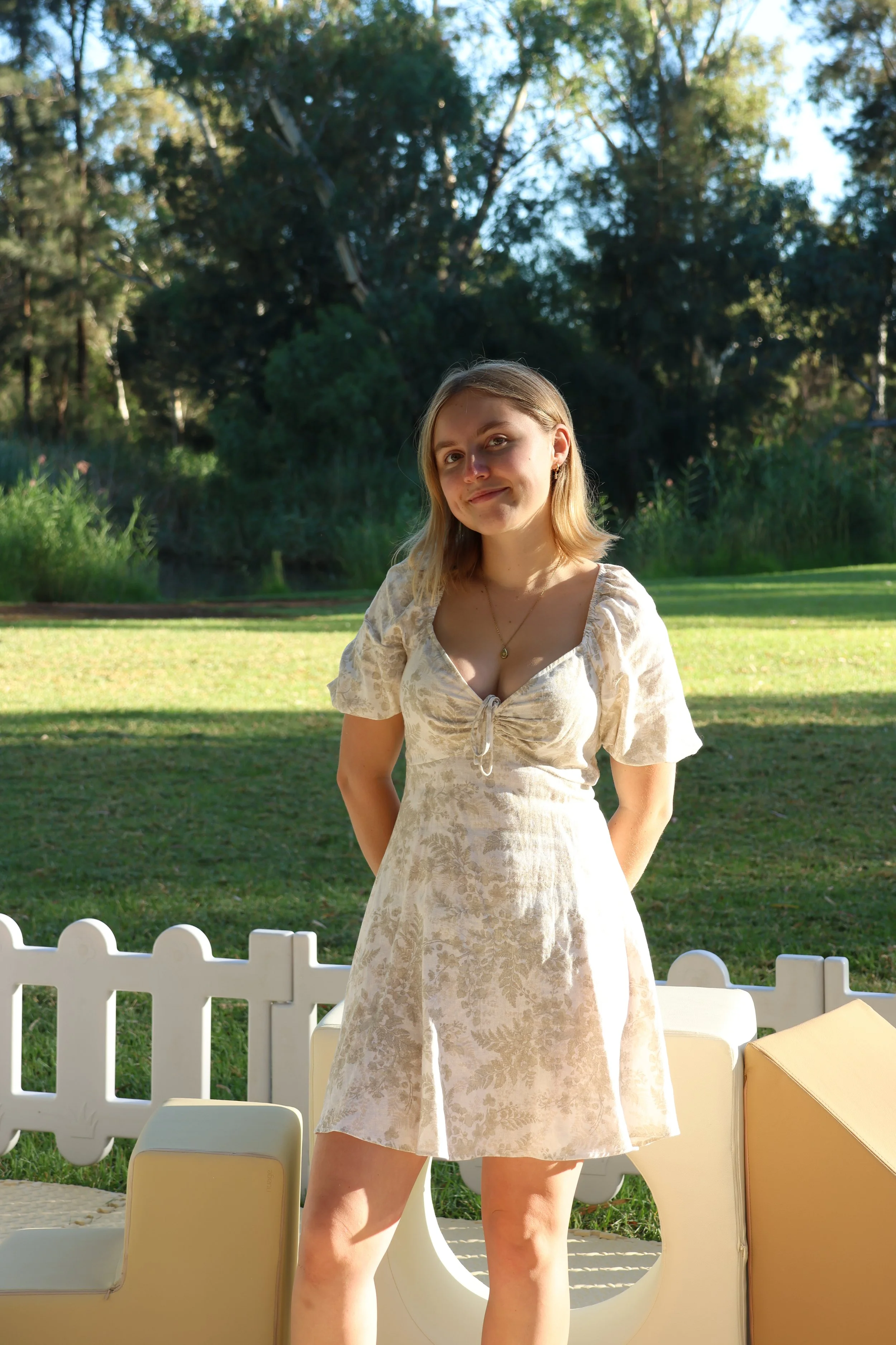 A young woman standing outside in a park or garden, wearing a light-colored dress, with a white picket fence and green trees in the background.