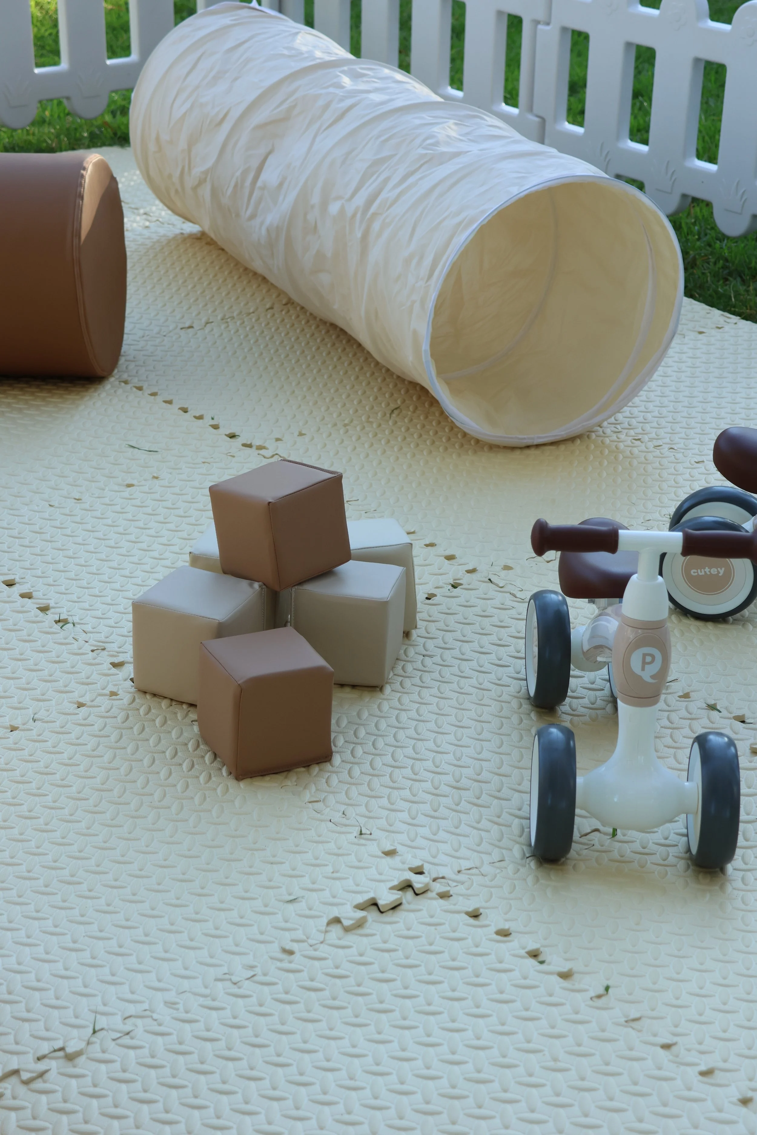 Outdoor play area with a white tunnel, toy scooter, and soft foam cubes on a textured yellow mat, surrounded by a white picket fence and green grass.