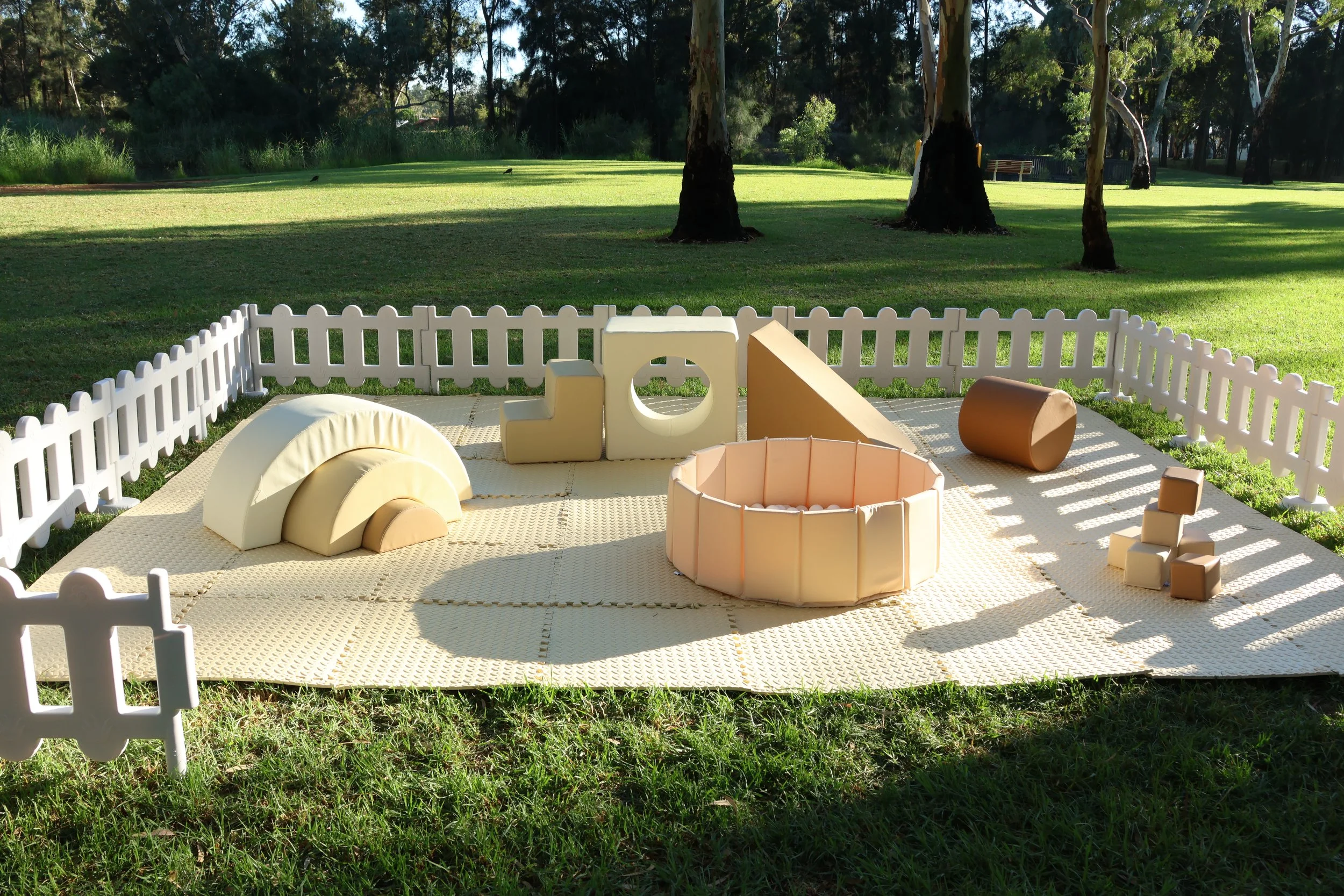 Children's outdoor play area on grass with foam climbing blocks, tunnel, and foam shapes, enclosed by a white picket fence, in a park-like setting with trees and sunlight.