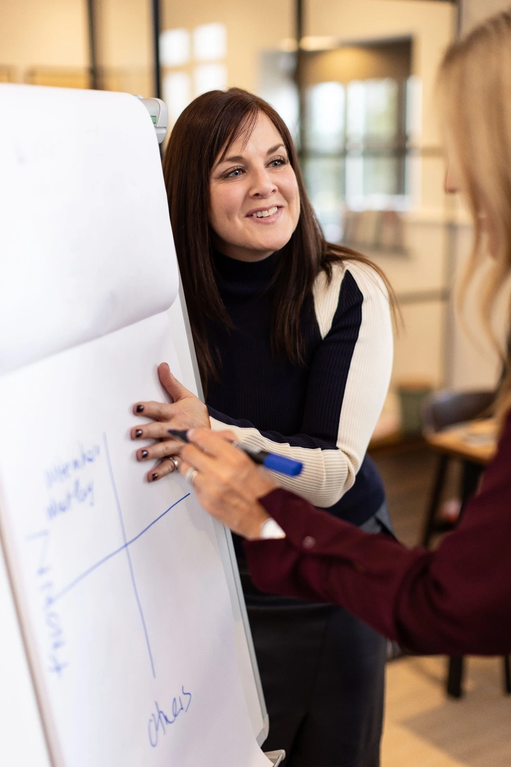 Lisa Truscott, a woman with long brown hair, wearing a black and cream striped sweater, smiling and engaging in a discussion with another person near a whiteboard in a well-lit room.