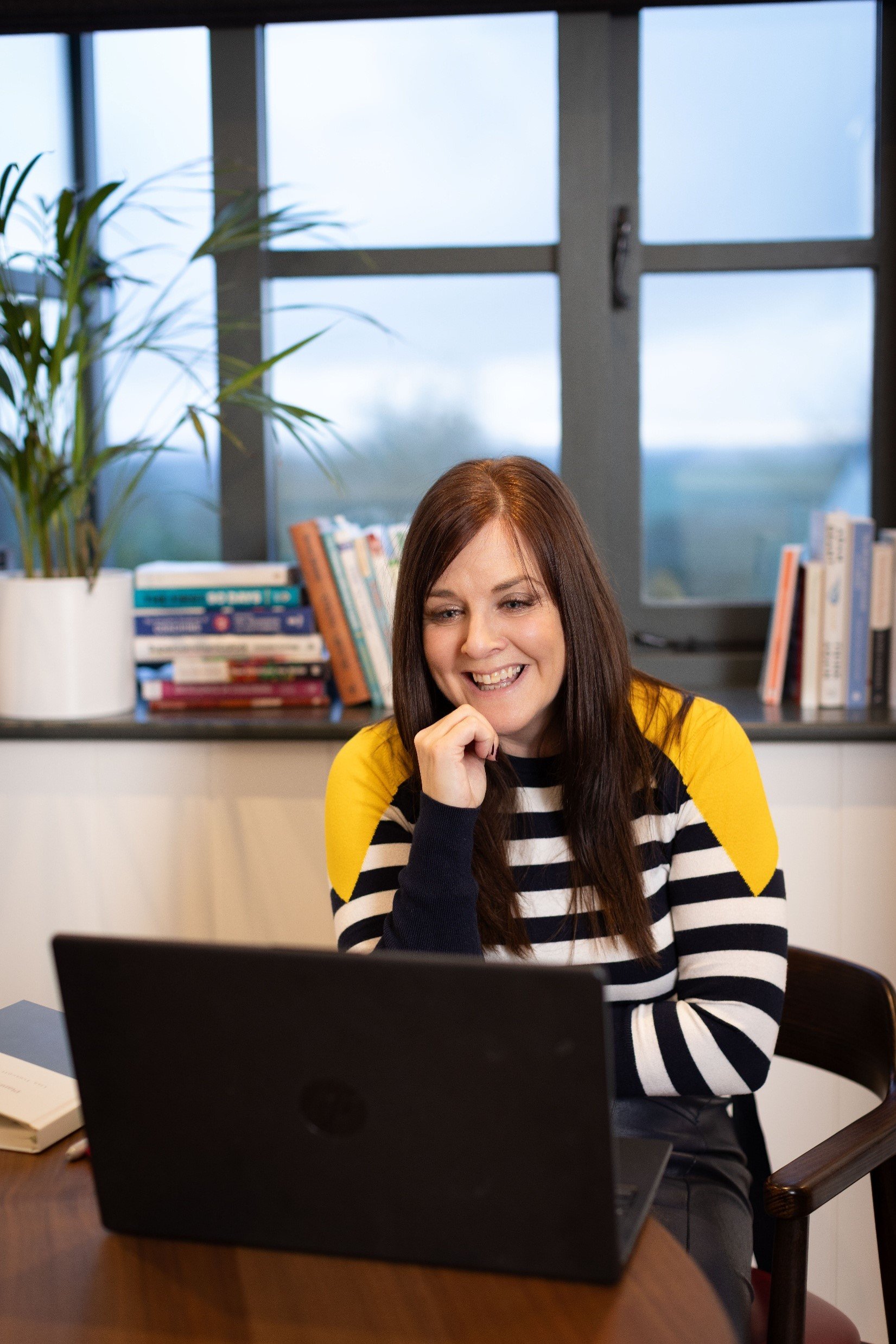 Lisa Truscott, a smiling woman with brown hair sitting at a desk in front of a laptop, with a bookshelf and window in the background.