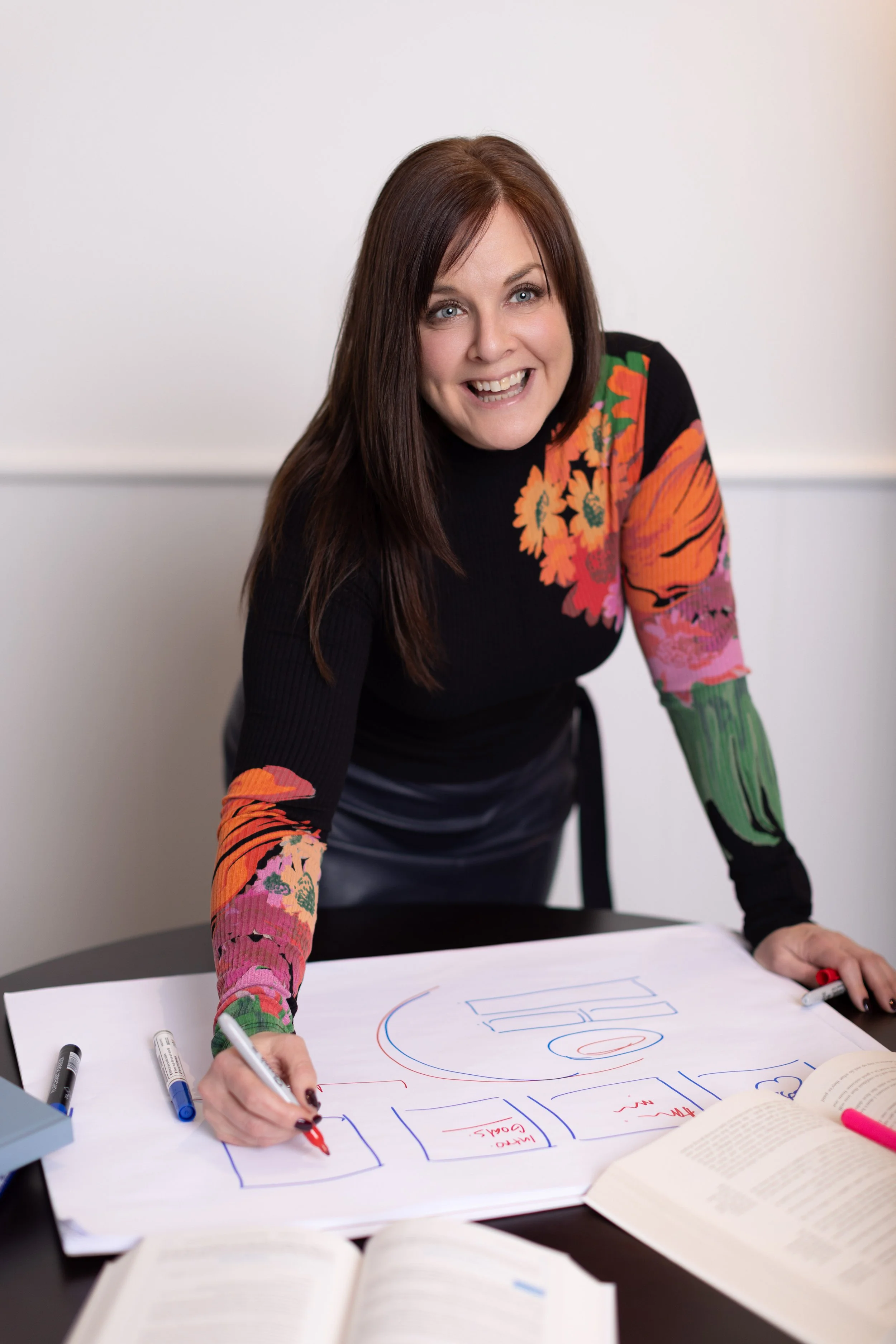 Lisa Truscott, a woman with long brown hair, wearing a black top with colorful floral sleeves, is standing over a table, drawing on a large sheet of paper with colorful markers. The table is scattered with books, markers, and a notebook.