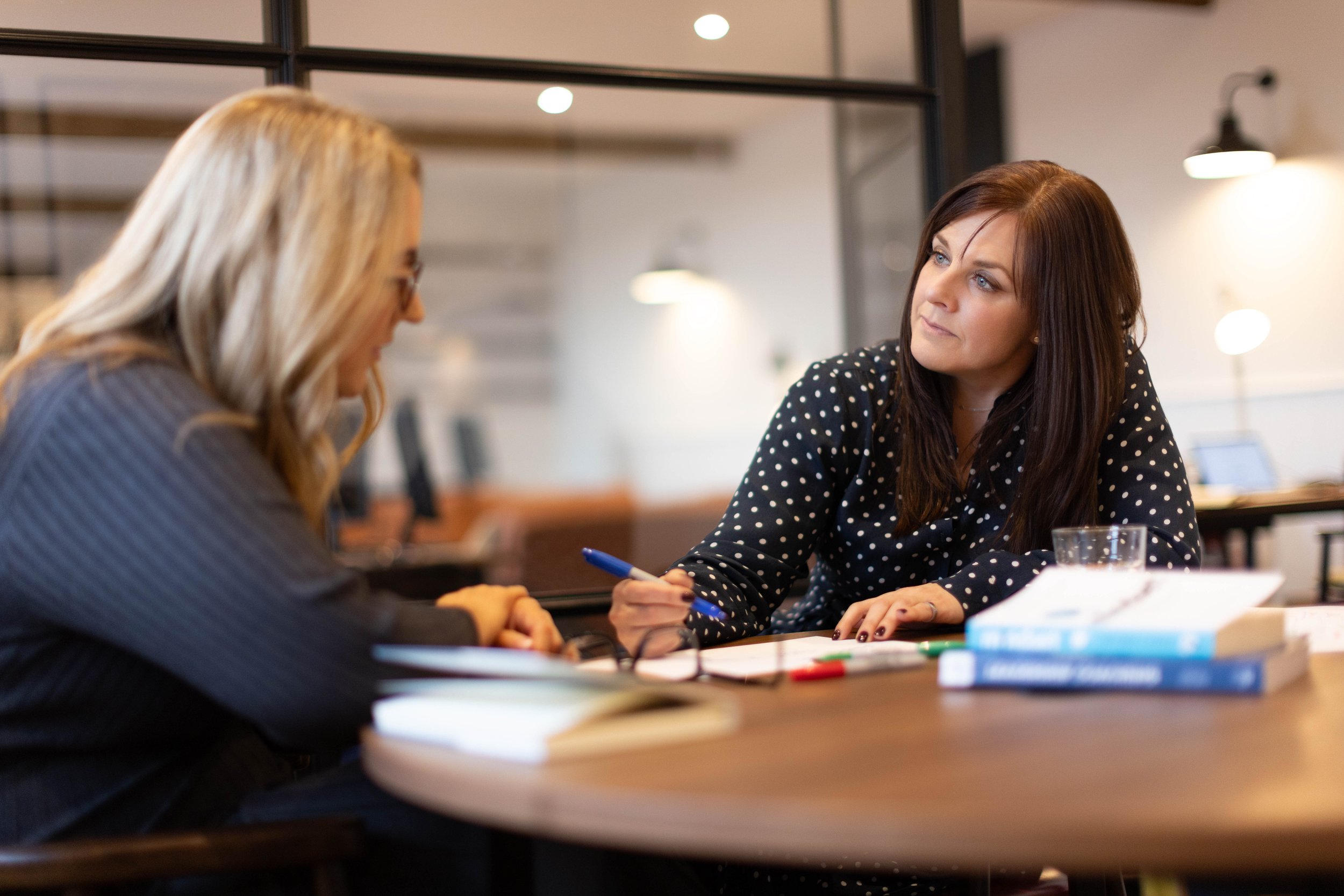 Two women are sitting across from each other at a table in a modern office or café setting. The woman on the right has long brown hair, blue eyes, and is wearing a black blouse with white polka dots. She is holding a pen and appears to be having a conversation with the woman on the left, who has blonde hair, wears glasses, and a gray striped blazer. There are notebooks and a glass of water on the table.