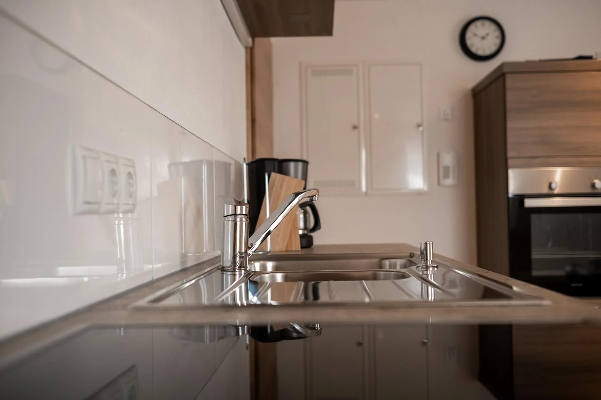Blick auf eine moderne Küchenspüle mit Wasserhahn, im Hintergrund Herd, Kaffeemaschine, Küchenwand mit Steckdosen und eine Uhr.