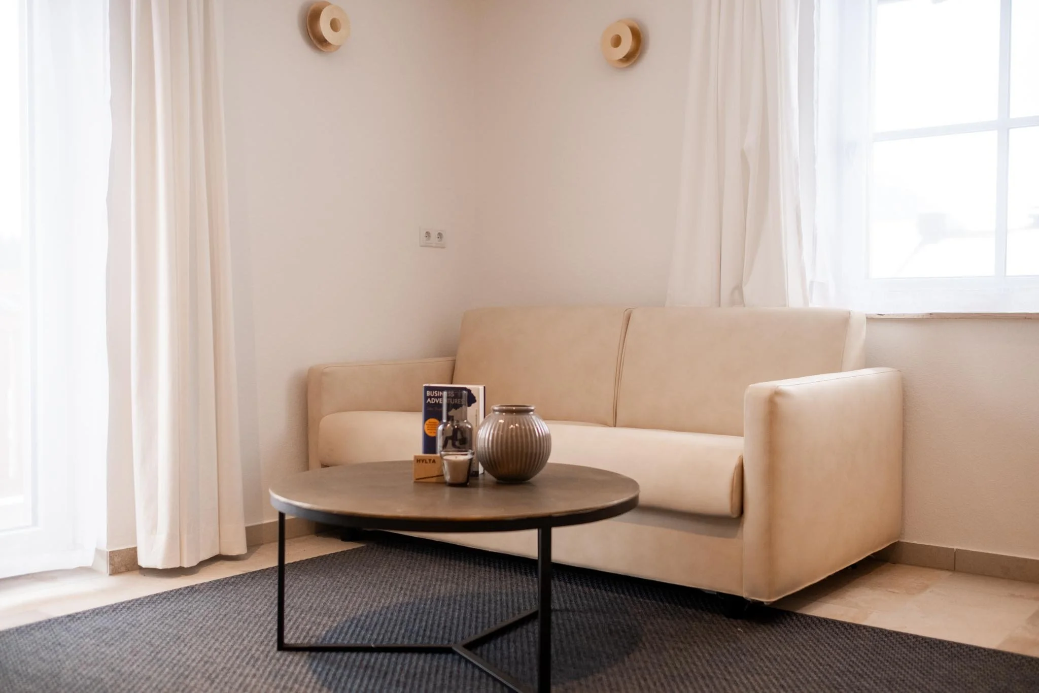 Gemütliches Wohnzimmer mit einer weißen Couch, einem kleinen runden Holztisch, Dekorationsgegenständen und einer großen Fensteröffnung mit weißen Vorhängen.