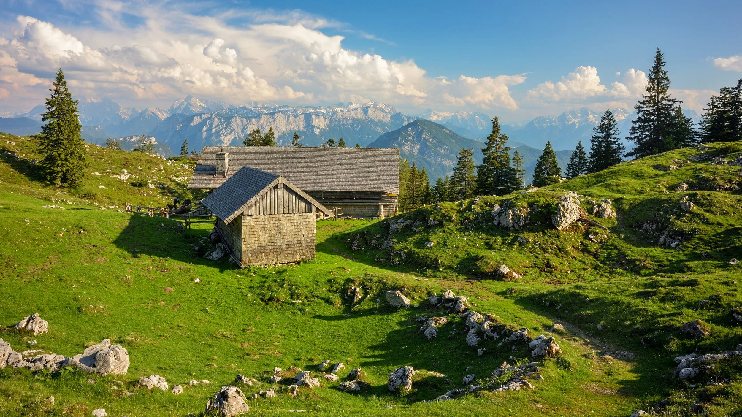 Grüne Wiese mit Steinhäusern inmitten von Bergen und Tannenbäumen, im Hintergrund schneebedeckte Berge unter einem Himmel mit Wolken.