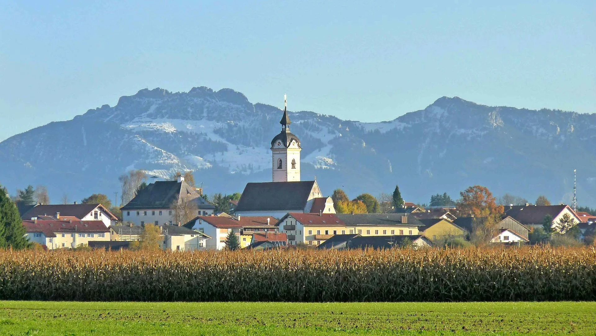 Blick auf ein Dorf mit Kirche vor Bergen, im Vordergrund ein Weizenfeld und Wiese, im Hintergrund Berge mit Schnee.