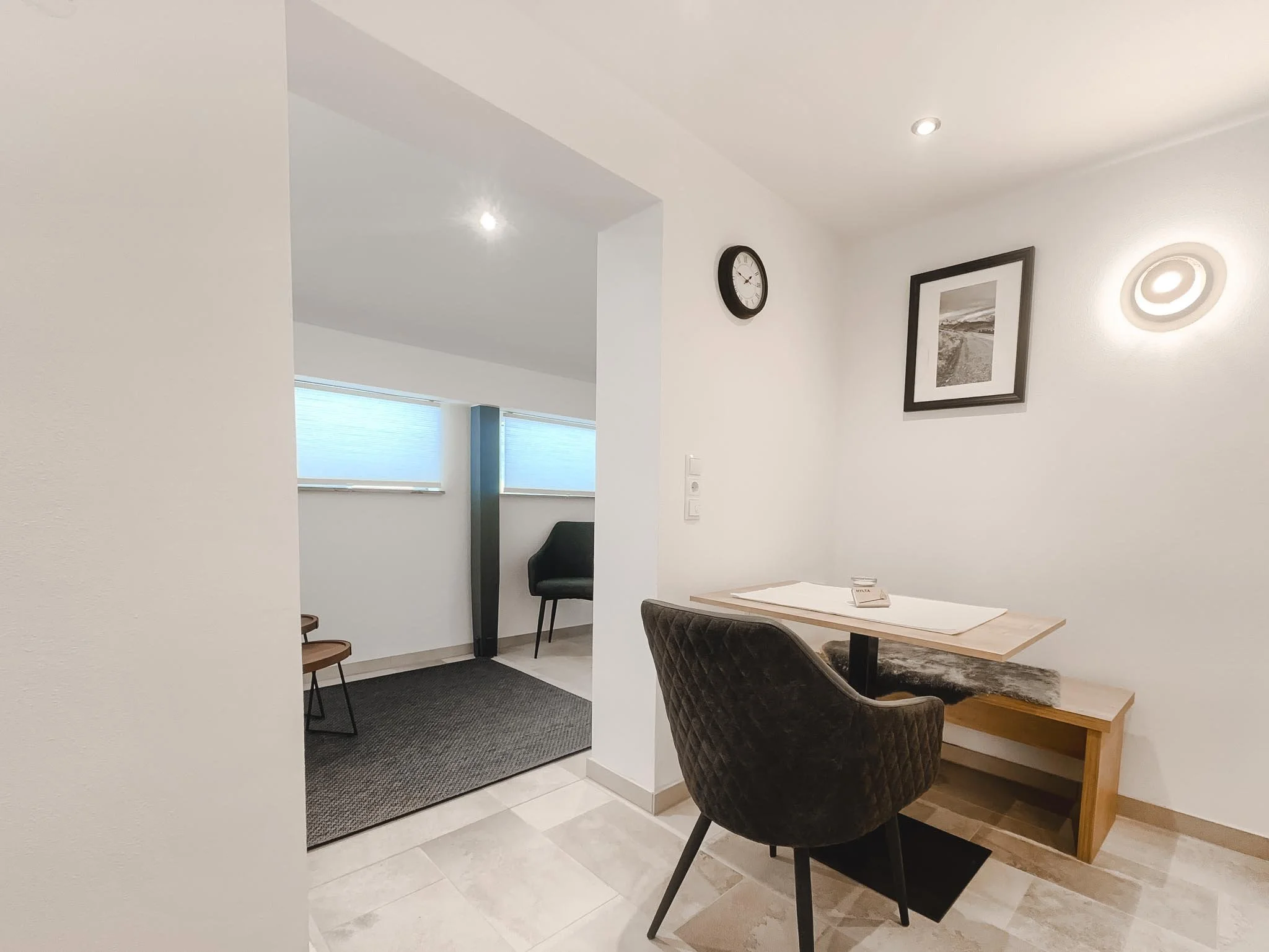 Ein kleines Büro mit einem schwarzen Sessel, einem Holztisch, einem Bilderrahmen an der Wand, einer Uhr und moderner Beleuchtung.