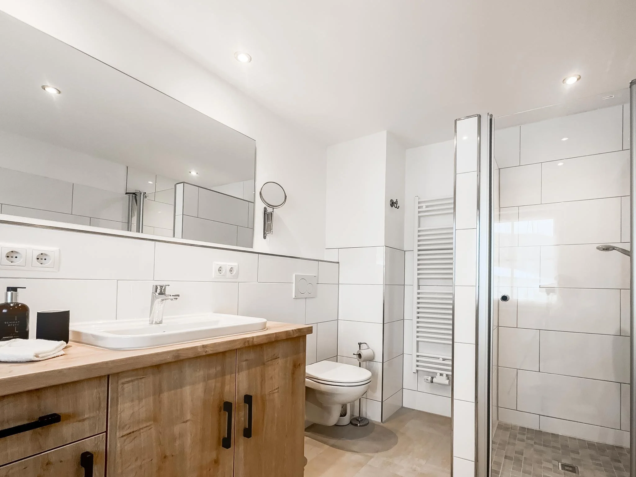 Modernes Badezimmer mit Holz-Waschtisch, großem Spiegel, Wandheizung, Toilette und begehbarer Dusche mit Glasabtrennung