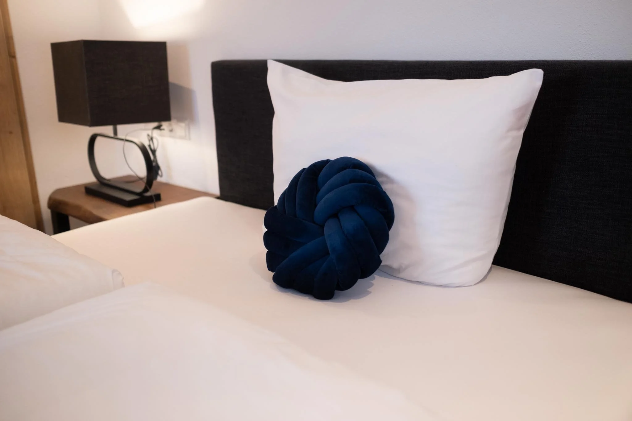 Ein Bett mit weißer Bettwäsche, weißem Kissen und einem dunkelblauen, knotigen Dekokissen. Im Hintergrund steht ein Nachttisch mit einer schwarzen Lampe und einem schwarzen Telefon.