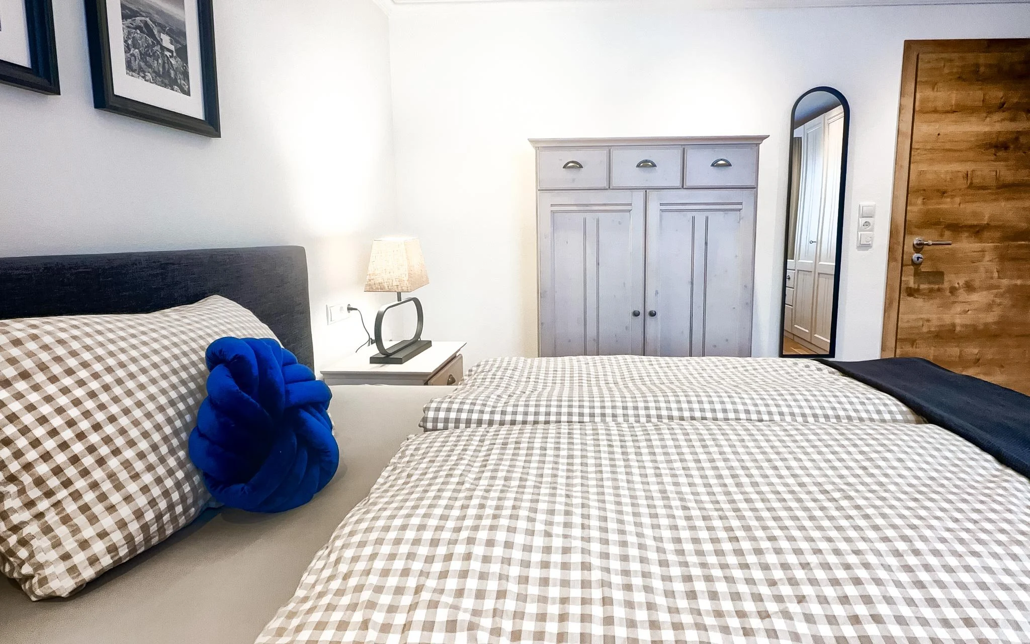 Schlafzimmer mit zwei Betten, Nachttisch mit Lampe, Kleiderschrank, Wandspiegel, Holztür, Bilder an der Wand, Kissen und Decken auf den Betten.