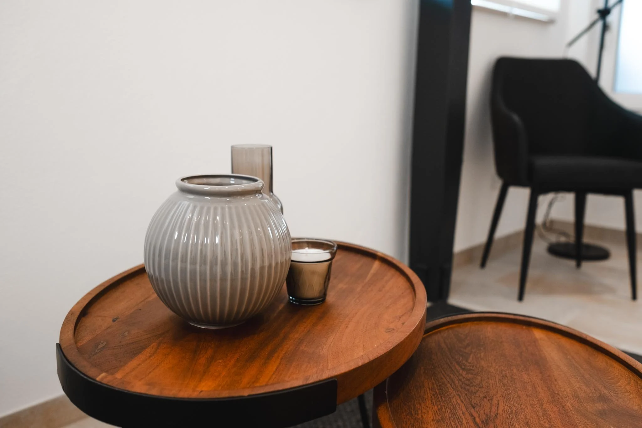Runde Holztablett mit weißen und schwarzen Keramikvasen und einer kleinen Kerzenwachskerze, im Hintergrund schwarzes Sessel in einem Modernen Wohnzimmer